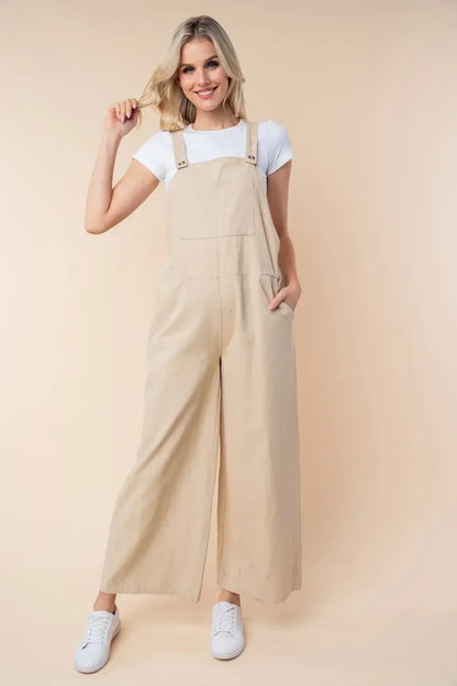 Woman wearing wide-leg beige overalls and white t-shirt, standing against a neutral background