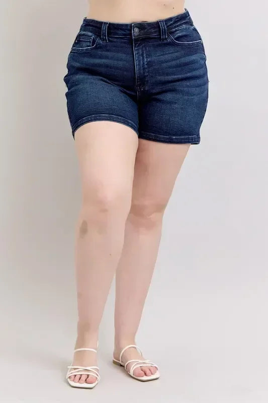 High-rise dark denim shorts styled with white strappy sandals on a neutral background.