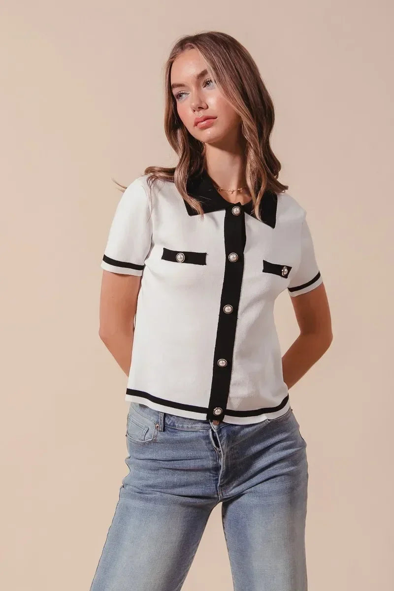 Woman wearing a short sleeve white knit top with black trim and pearl buttons, paired with jeans
