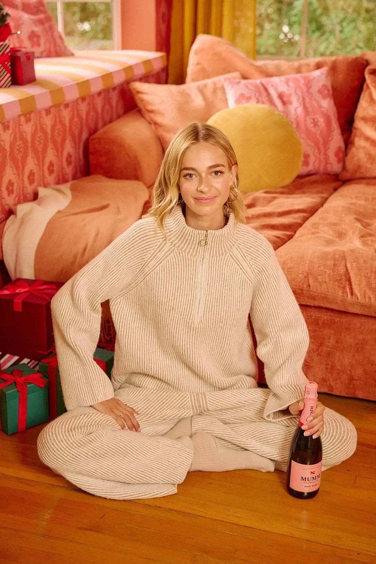 Woman in a cream ribbed knit lounge set sitting by peach sofa with wrapped gifts and rosé bottle