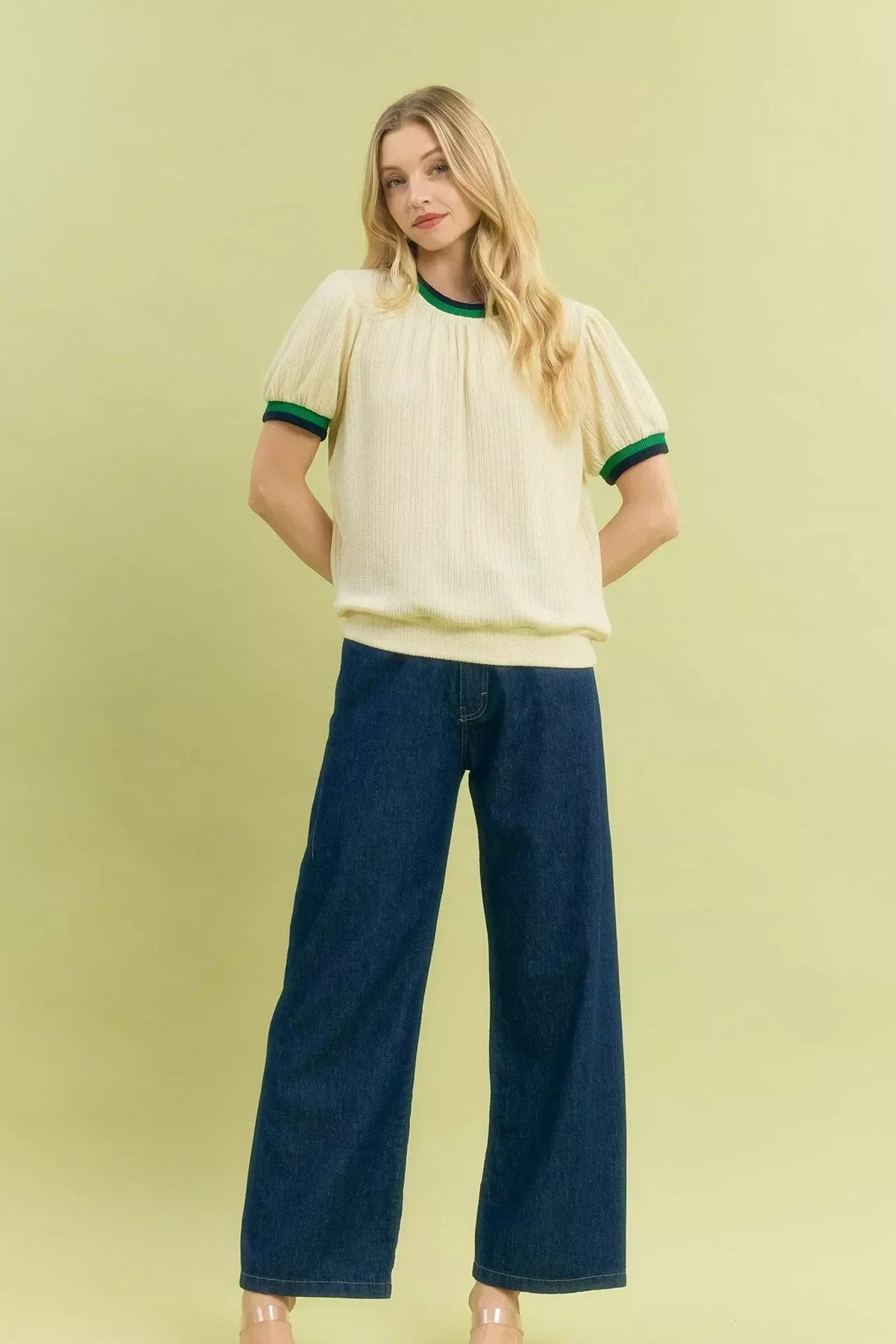 Woman wearing a short sleeve cream knit top with green trim and wide-leg dark blue jeans.
