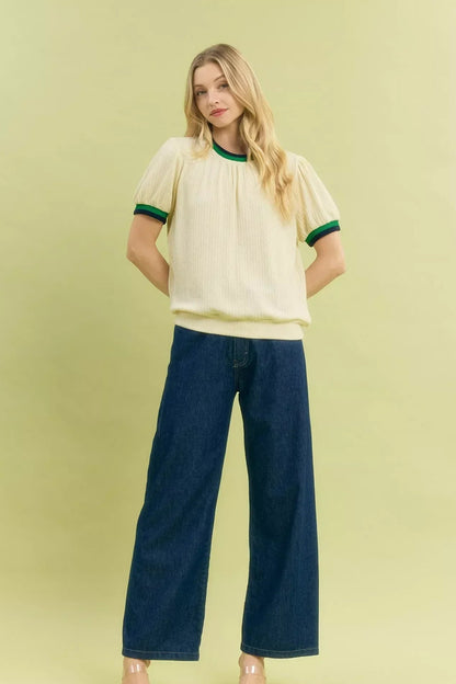 Woman wearing a short sleeve cream knit top with green trim and wide-leg dark blue jeans.