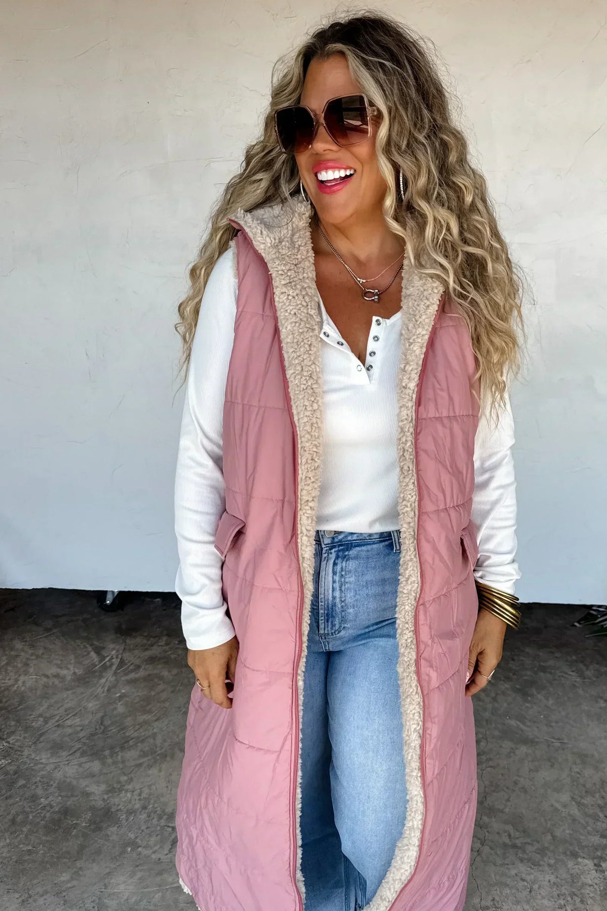 Woman in a pink quilted long vest with sherpa lining, white henley top, and blue jeans