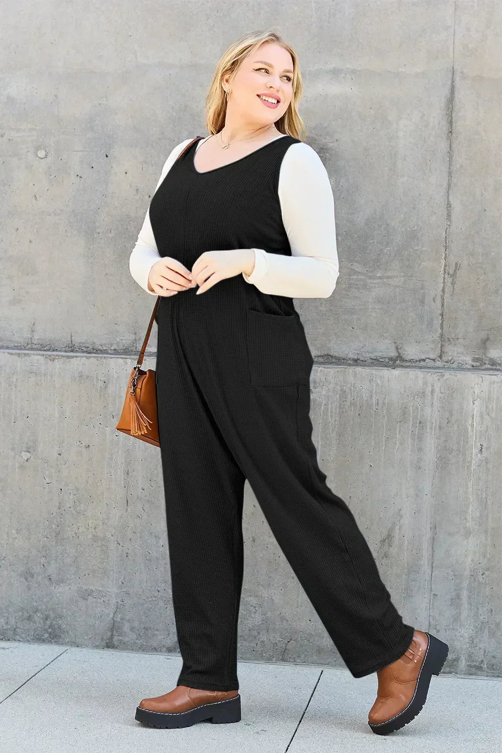 Woman wearing a black sleeveless jumpsuit over a cream long-sleeve top, brown shoes, and crossbody bag.