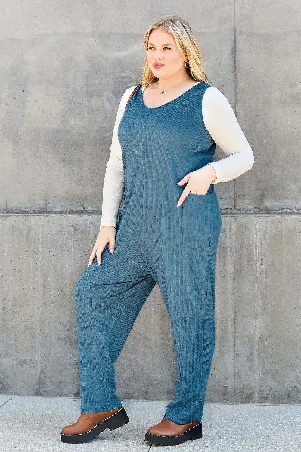 Woman wearing a relaxed blue jumpsuit over a long sleeve top with brown platform shoes, standing against a concrete wall