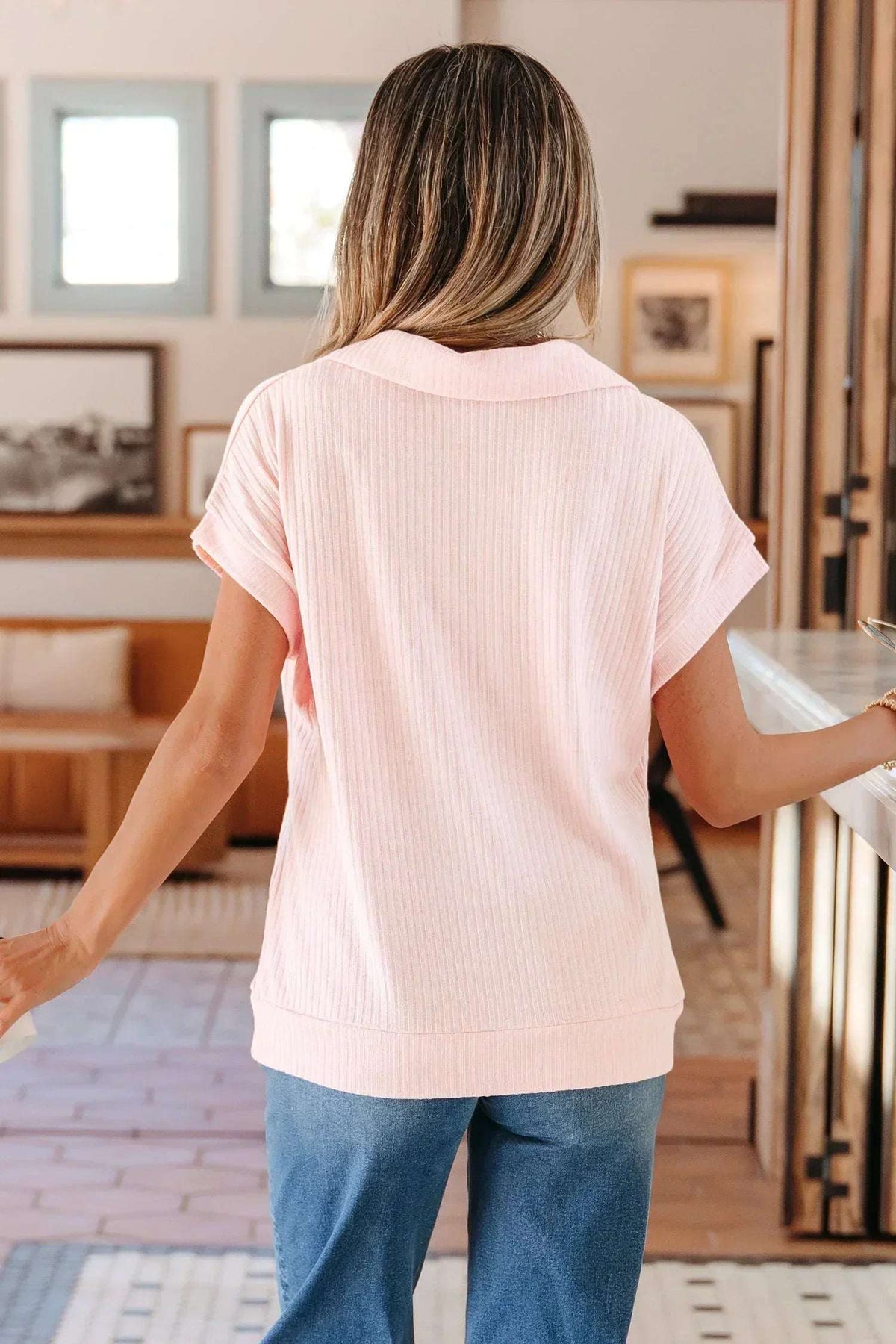 Woman in a light pink short-sleeve knit top and blue jeans standing indoors