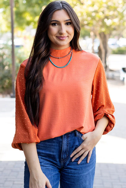 Woman in textured orange blouse with long sleeves and high neck, paired with blue jeans outdoors.