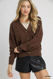 Woman wearing a brown cable knit cardigan with button front and black faux leather shorts.