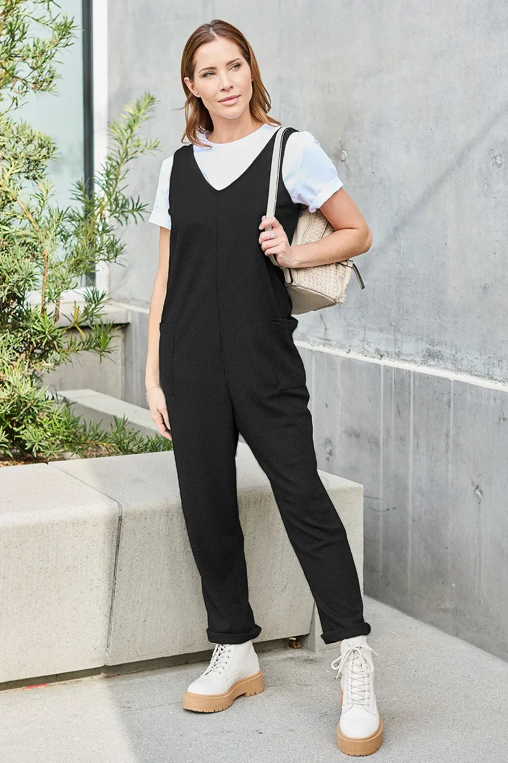Woman wearing a black sleeveless jumpsuit over a white t-shirt with chunky boots, carrying a beige bag, standing outdoors by a concrete wall.