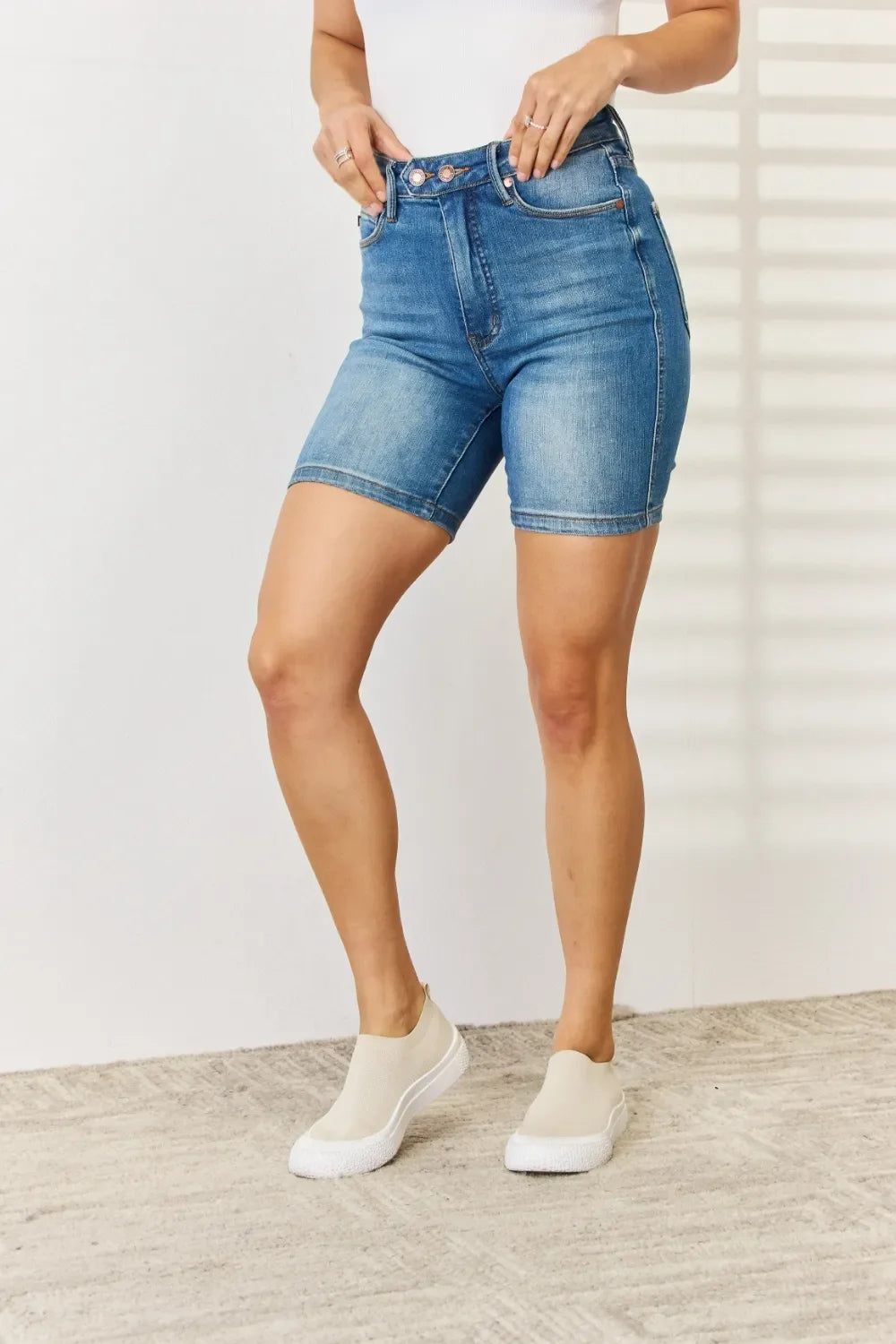 Woman wearing high-waisted denim Bermuda shorts and slip-on sneakers, standing indoors