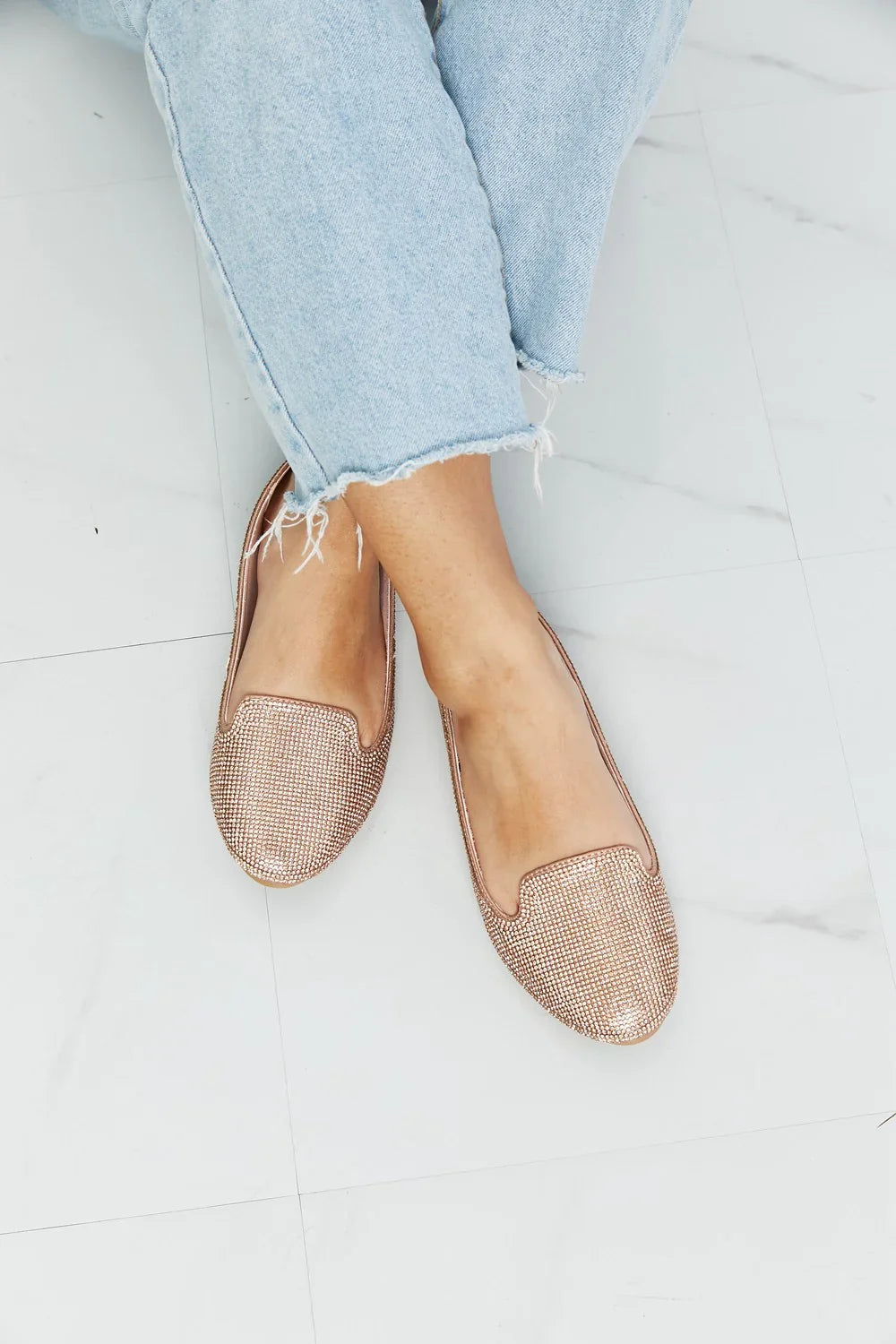 Rose gold rhinestone embellished loafers paired with light wash frayed hem jeans on tile floor