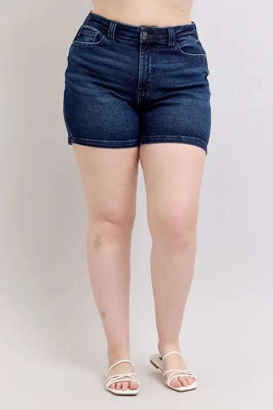 Woman wearing high-waisted dark denim shorts and white sandals against a neutral background