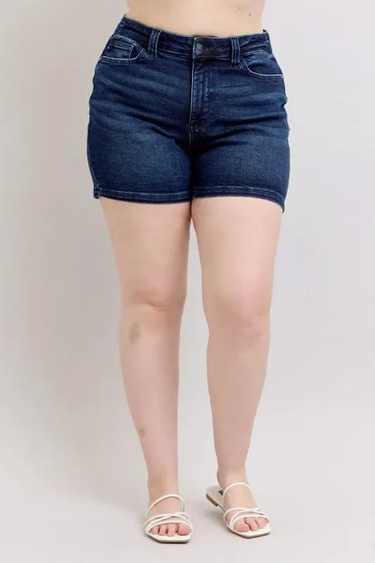 Woman wearing high-waisted dark denim shorts and white sandals against a neutral background