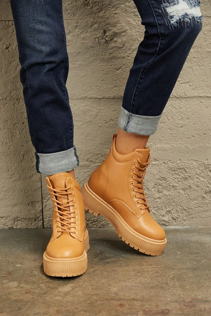 Tan lace-up platform boots styled with rolled dark denim jeans against a concrete wall