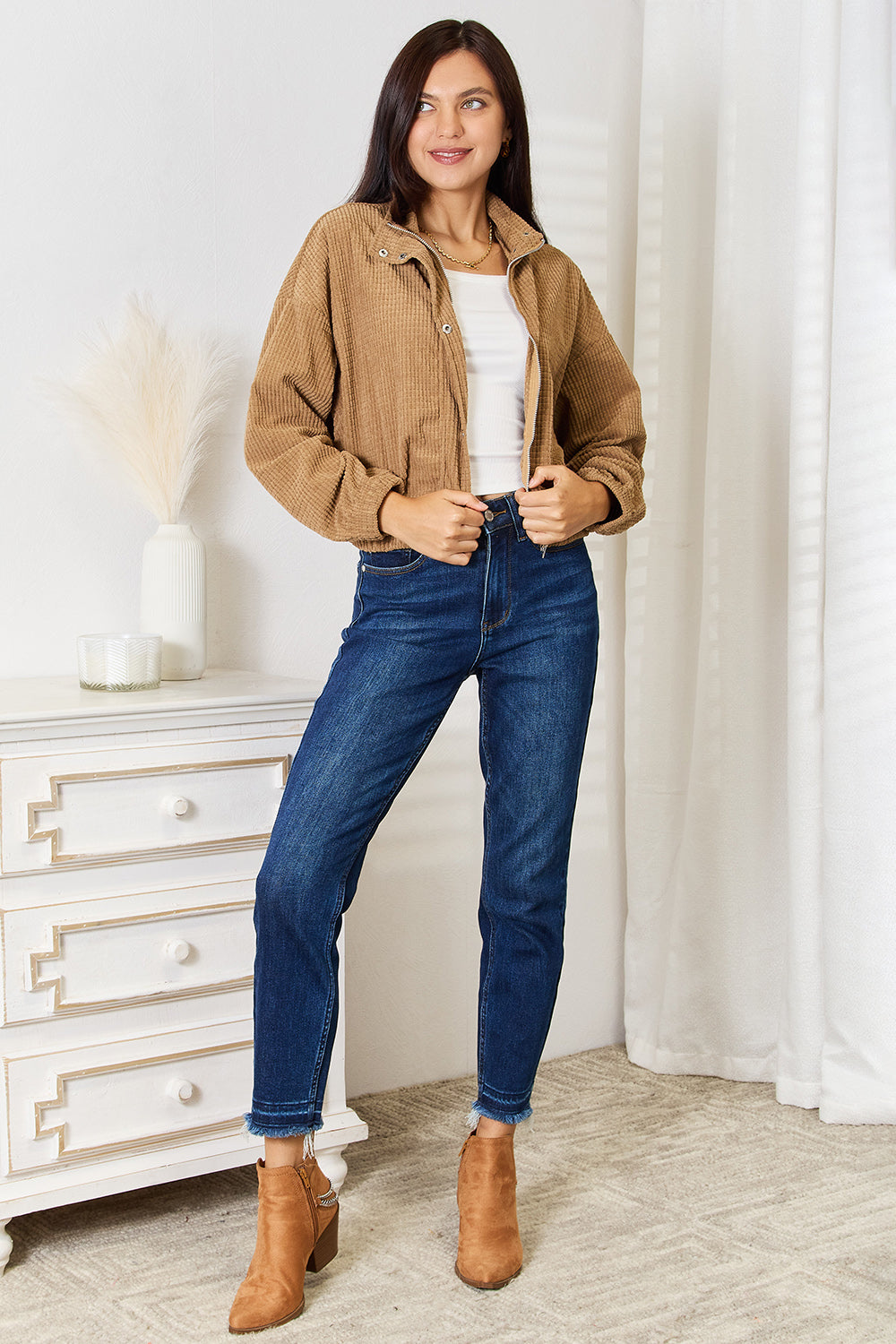 Woman wearing a brown Long Sleeve Dropped Shoulder Jacket, white top, blue jeans, and brown boots in a room with a white dresser and curtains.