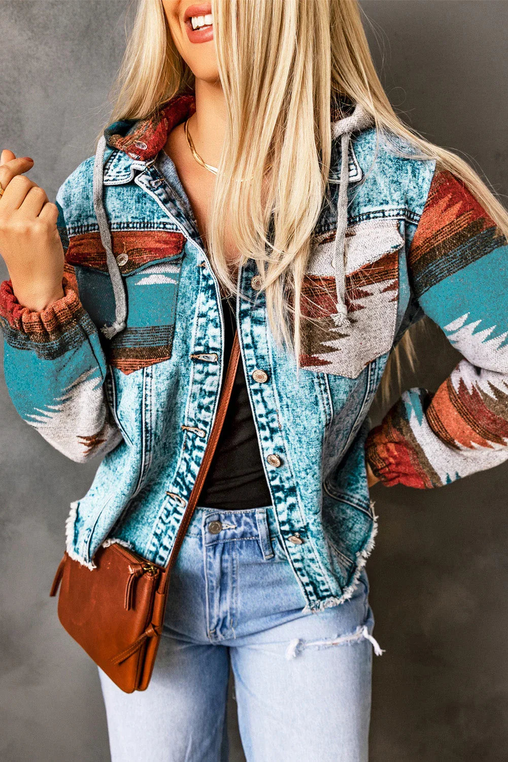 Woman in distressed denim jacket with colorful tribal print sleeves, light wash jeans, and brown crossbody bag