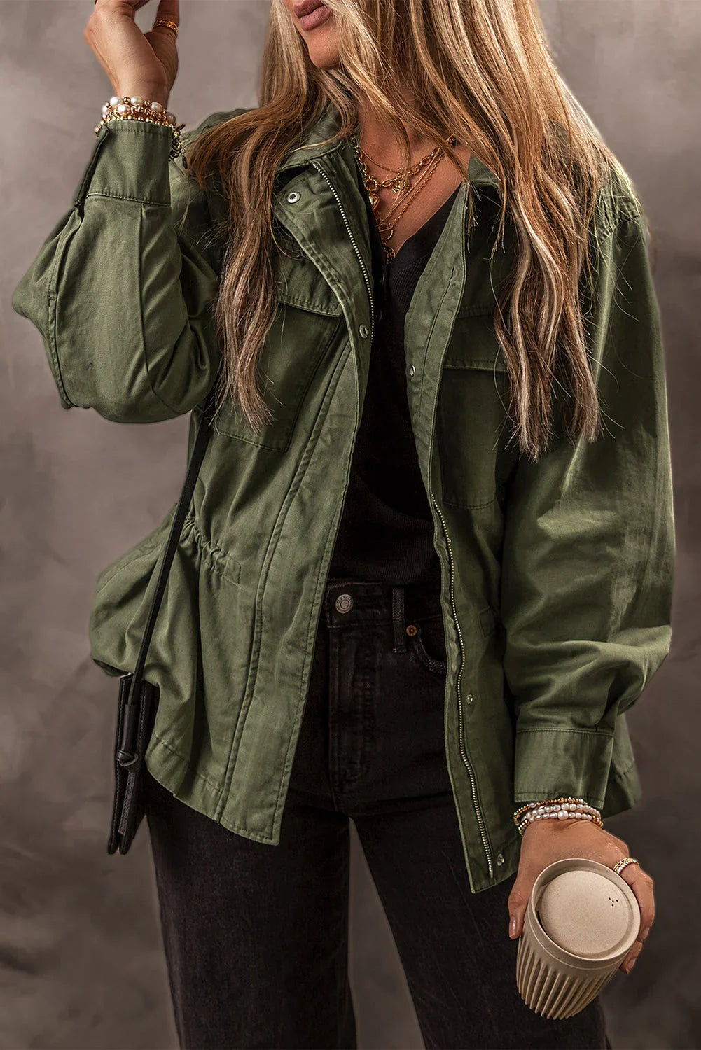 Woman wearing an olive green utility jacket, black top and jeans, holding a coffee cup