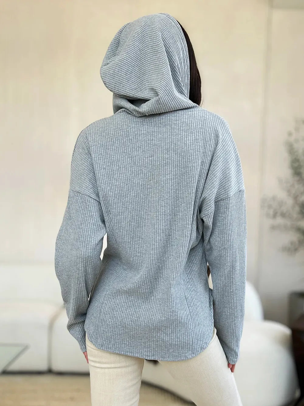 Woman wearing light grey ribbed hooded sweater and cream pants, shown from the back indoors