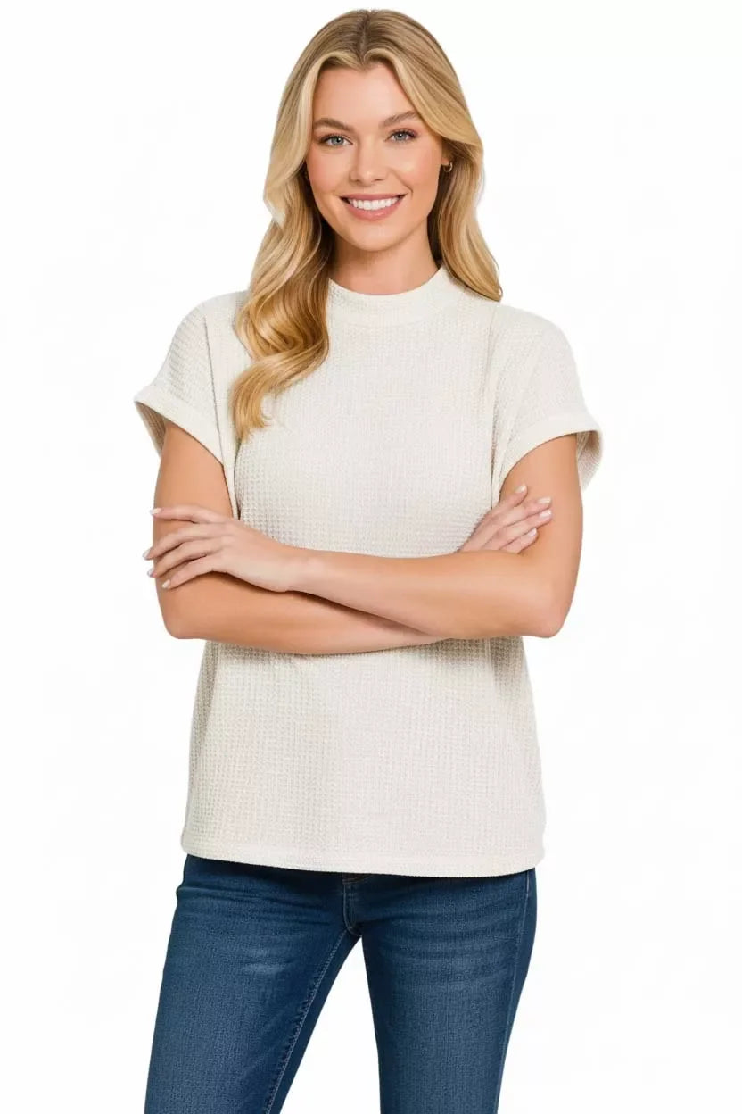 Woman wearing a textured cream short sleeve top and blue jeans, standing against white background