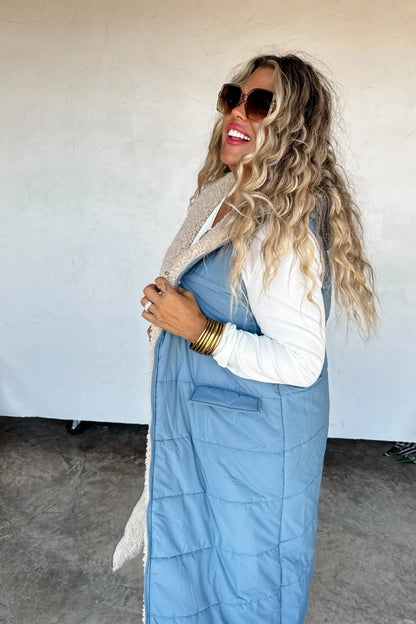 Woman in blue quilted long vest with faux shearling lining, wearing sunglasses and gold bangles