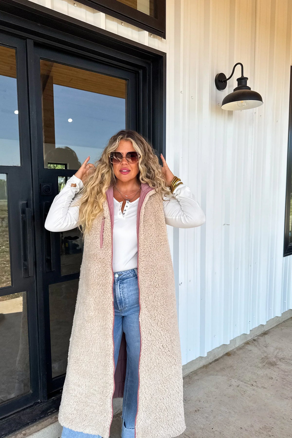 Woman in long sherpa vest, white top, and jeans standing outdoors by glass doors