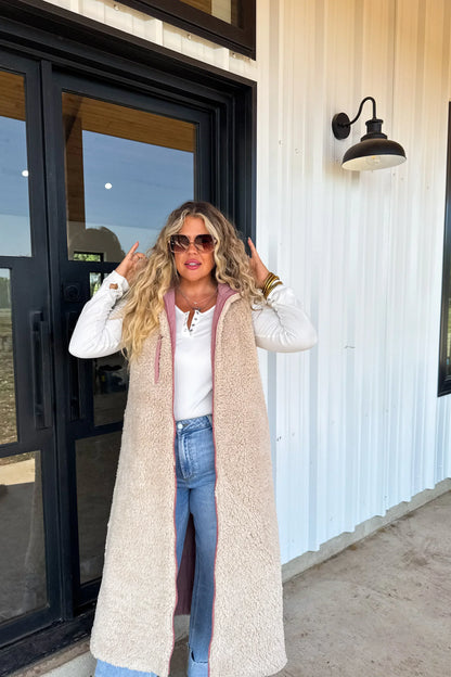 Woman in long sherpa vest, white top, and jeans standing outdoors by glass doors