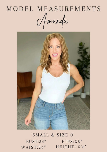 Woman modeling casual summer outfit with white tank top and high-waisted denim shorts indoors