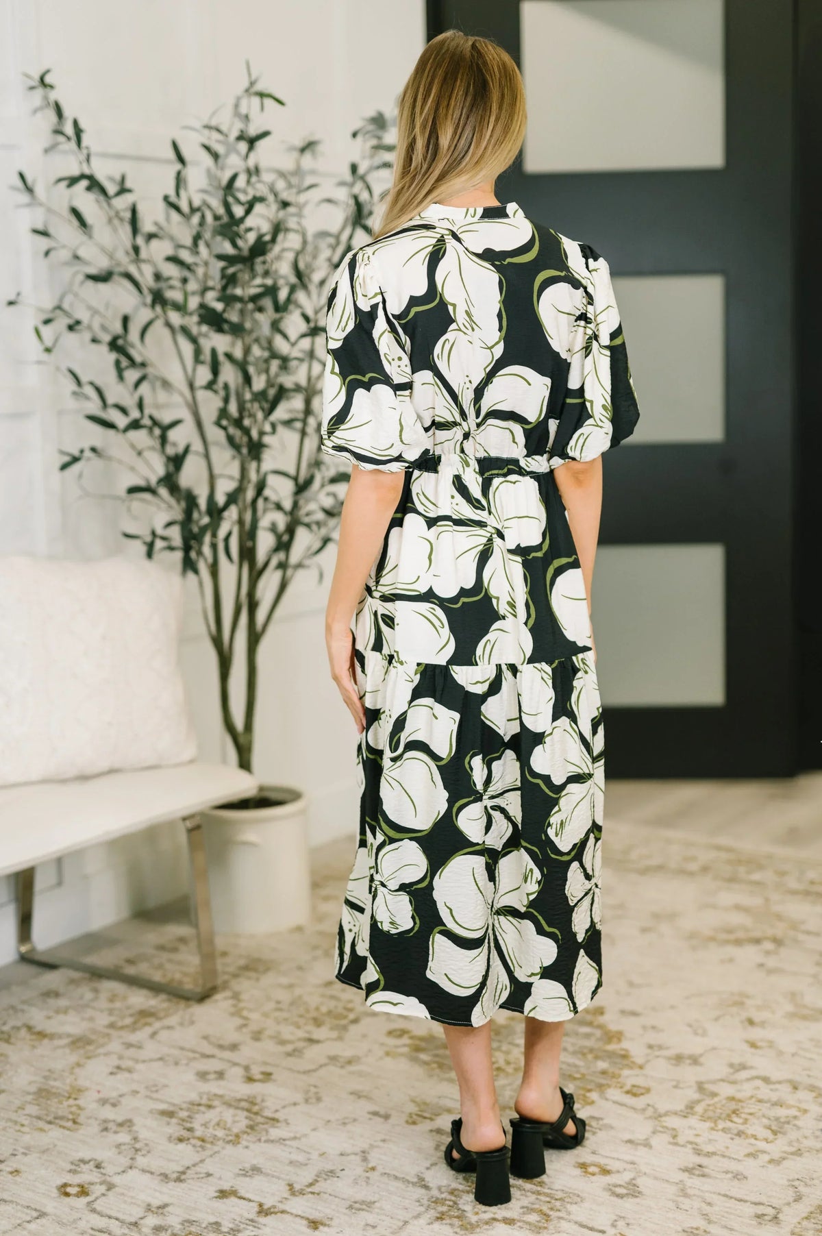 Woman wearing a black floral puff sleeve midi dress, standing indoors on a patterned rug.