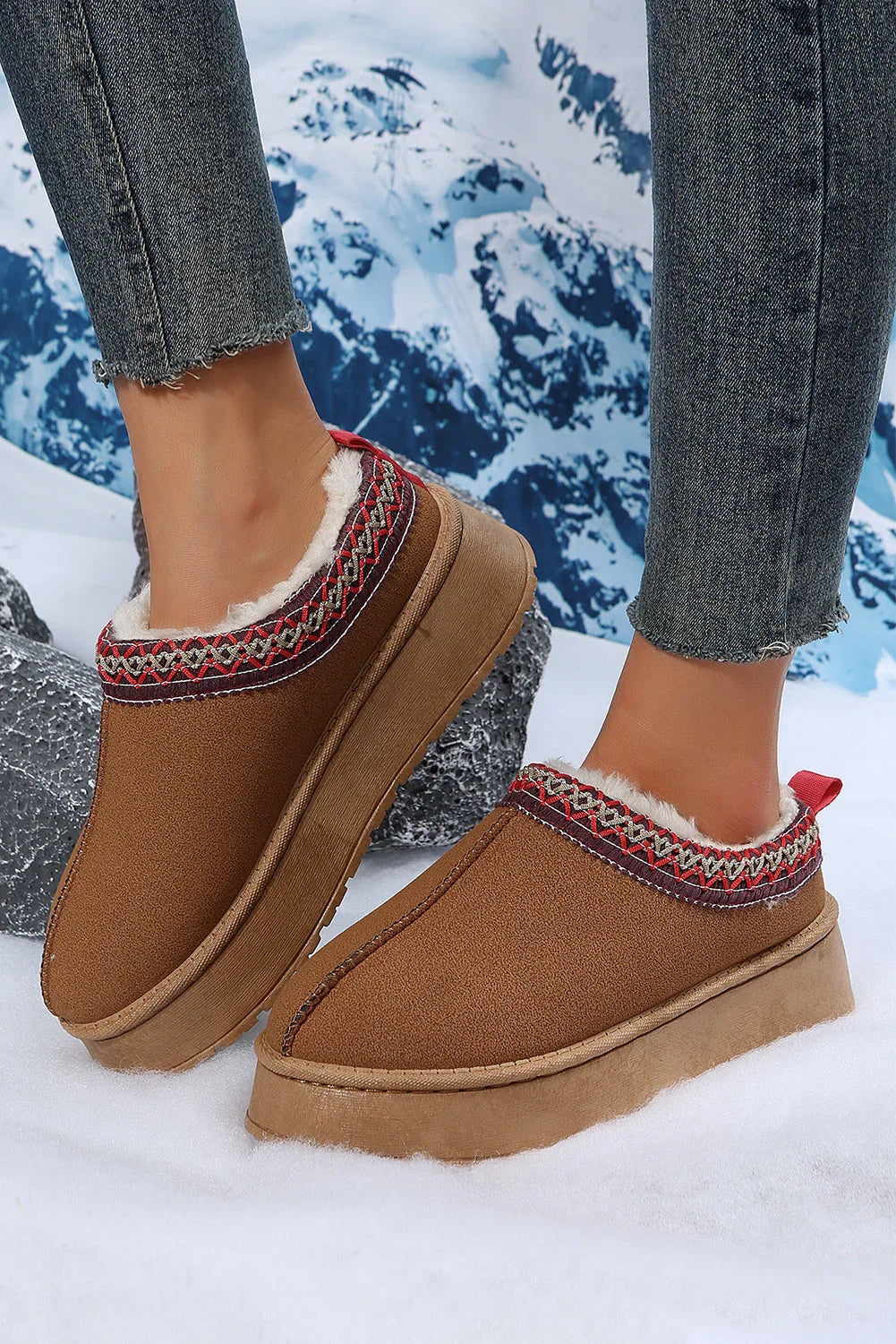Cozy brown shearling-lined slip-on slippers with patterned trim, worn with jeans on snow.