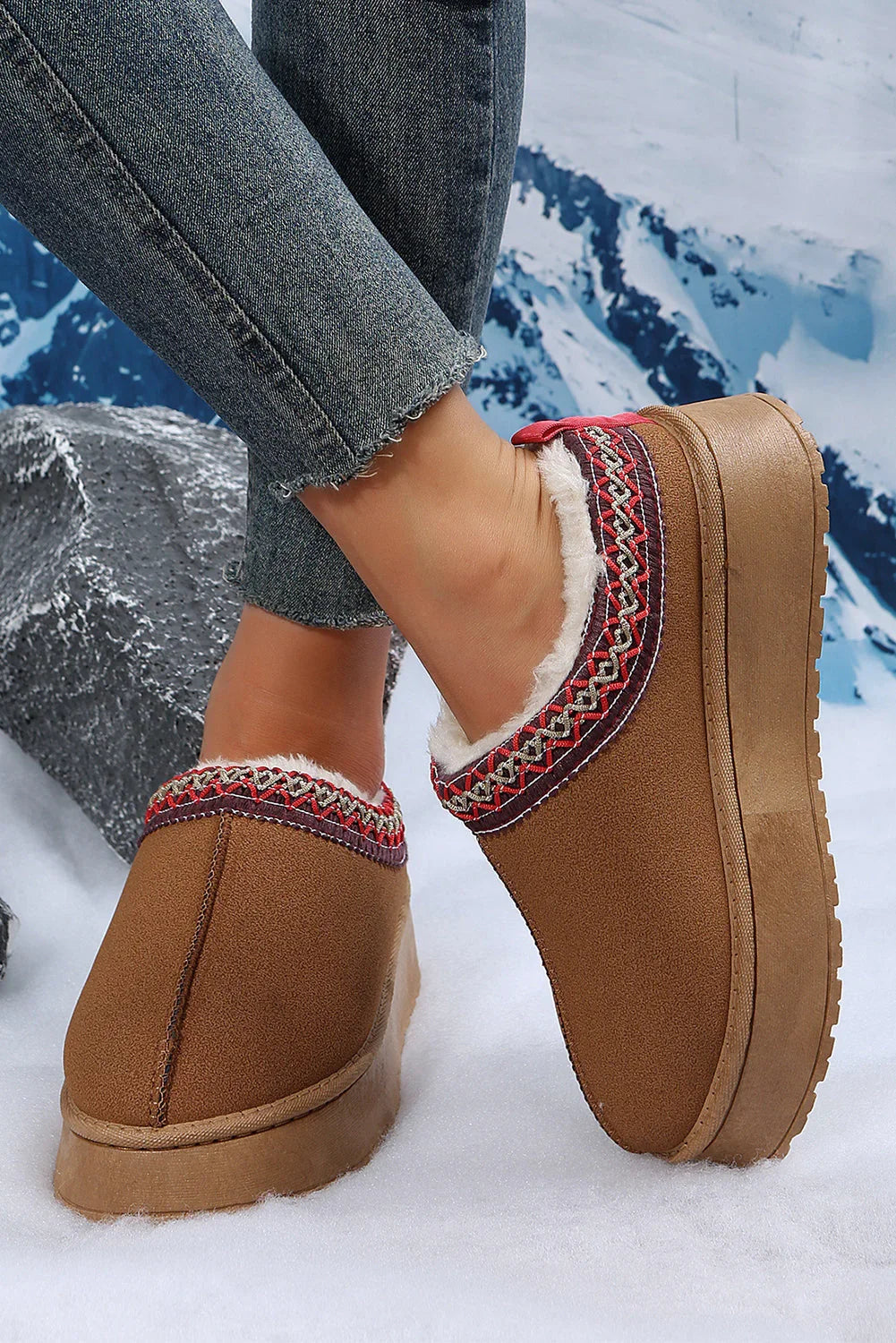 Brown suede platform slip-on slippers with faux fur lining and patterned trim, worn with jeans on a snowy setting.