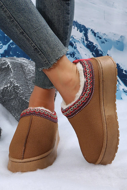 Brown suede platform slip-on slippers with faux fur lining and patterned trim, worn with jeans on a snowy setting.