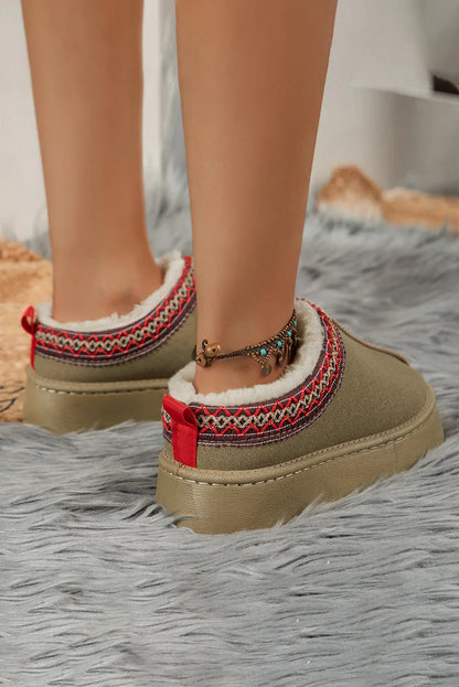 Cozy taupe slip-on slippers with red and patterned trim, faux fur lining, on faux fur rug.