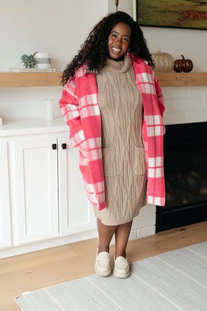 Woman in tan cable knit turtleneck sweater dress with pink plaid coat, standing indoors