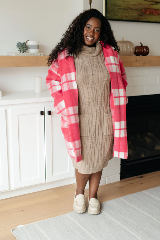 Woman in tan cable knit turtleneck sweater dress with pink plaid coat, standing indoors