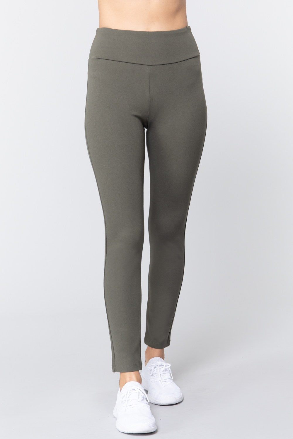 Woman wearing olive green waist band long ponte pants with white sneakers on a white studio background.
