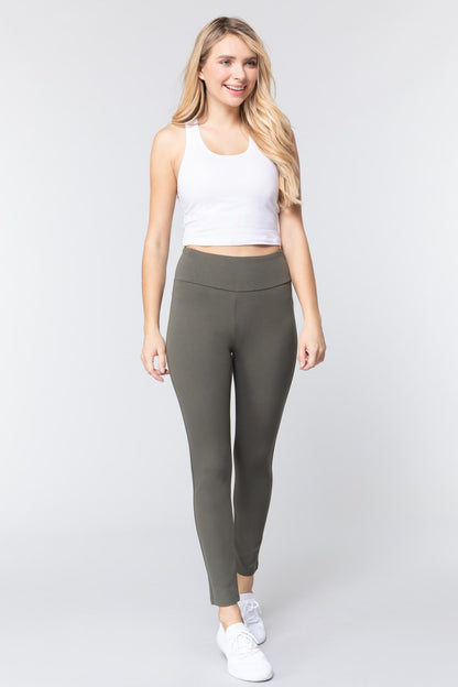Woman wearing olive green waist band long ponte pants and a white tank top with white sneakers on a white studio background.