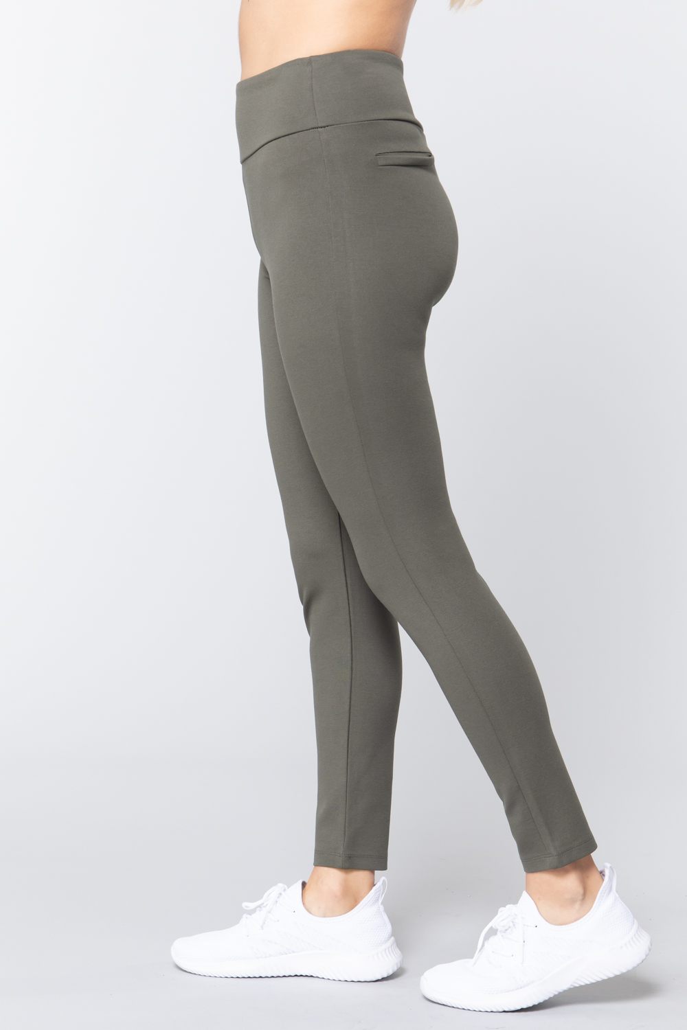 Woman wearing olive green waist band long ponte pants with back slit pockets with white sneakers on a white studio background.