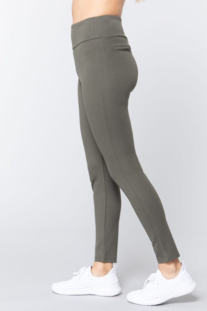 Woman wearing olive green waist band long ponte pants with back slit pockets with white sneakers on a white studio background.