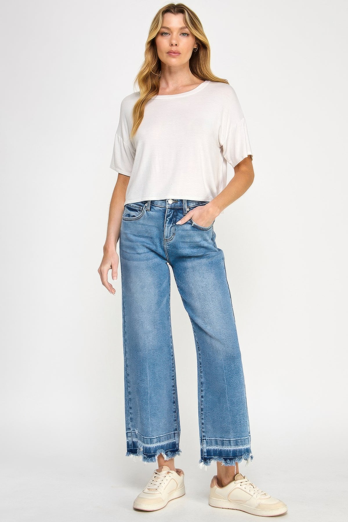 Woman wearing a white top and light Tummy Control Cropped Wide Leg Jeans with a frayed hem with white sneakers on a white background