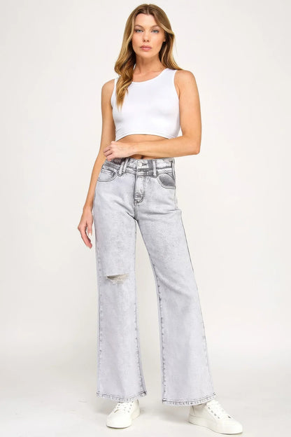 Woman in white crop tank top and light wash wide-leg jeans with knee rip, standing studio shot