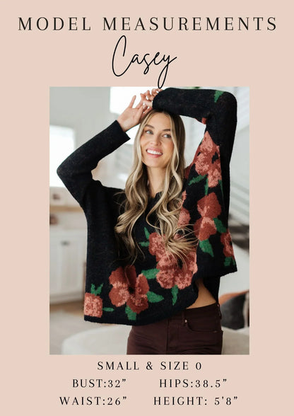 Woman modeling a loose black sweater with red floral pattern, standing in a bright home setting.