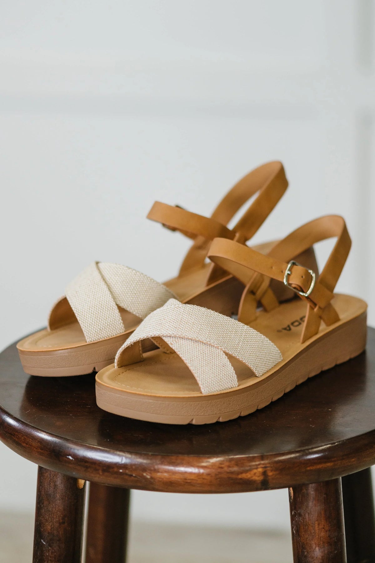 Cream nubuck wedge sandals with tan ankle straps and crisscross fabric detail on a stool