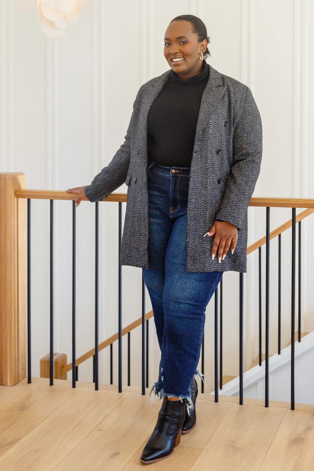 Woman wearing a black button-down blazer jacket, black turtleneck, blue jeans, and ankle boots standing indoors by a wooden railing.