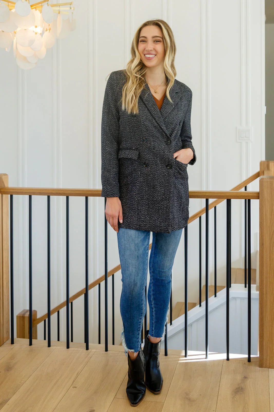 Woman wearing a black button-down blazer jacket, blue skinny jeans, and black ankle boots indoors.