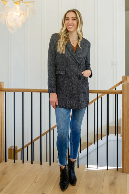 Woman wearing a black button-down blazer jacket, blue skinny jeans, and black ankle boots indoors.