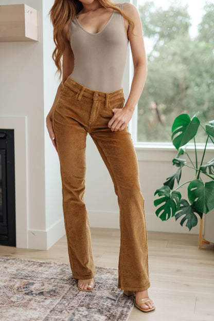 Woman wearing a beige tank top and brown boot cut corduroy pants in a room with a fireplace and plant.