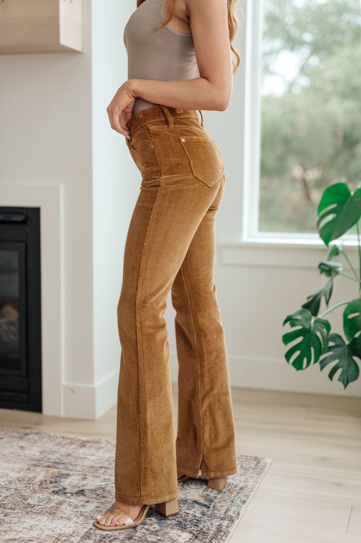 Side view of a woman wearing a beige tank top and brown boot cut corduroy pants in a room with a fireplace and plant.