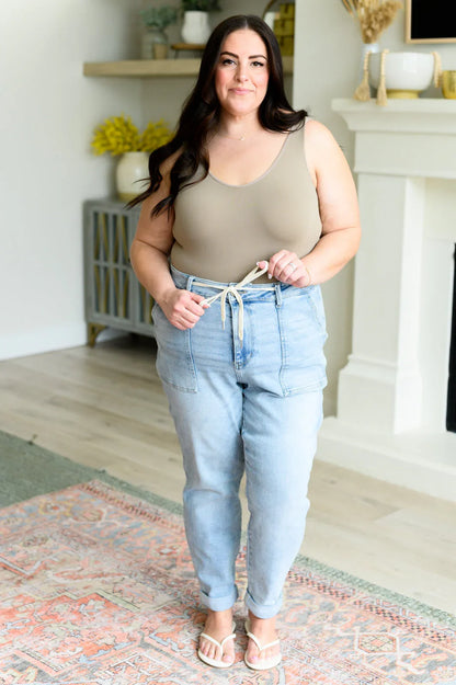 Woman in a beige tank top and light blue high-waisted jeans with drawstring, standing indoors.