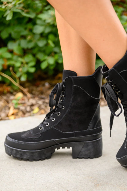Black lace-up suede boots with chunky heel and side stitching, worn outdoors on pavement
