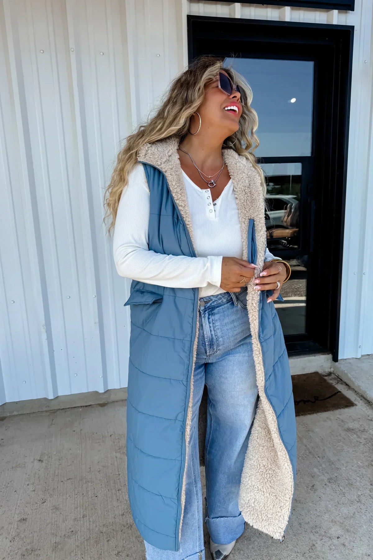 Woman wearing a long blue quilted sherpa-lined vest, white henley top, and light wash jeans